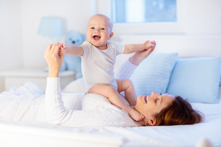 A mother, laying on a bed, holds her smiling baby up on her chest.