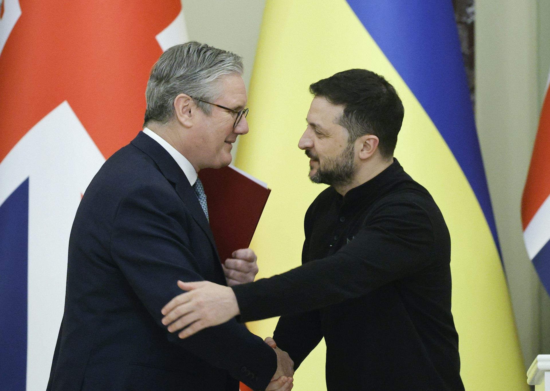Starmer and Zelensky shaking hands at a recent meeting, with UK and Ukraine flags in the background.