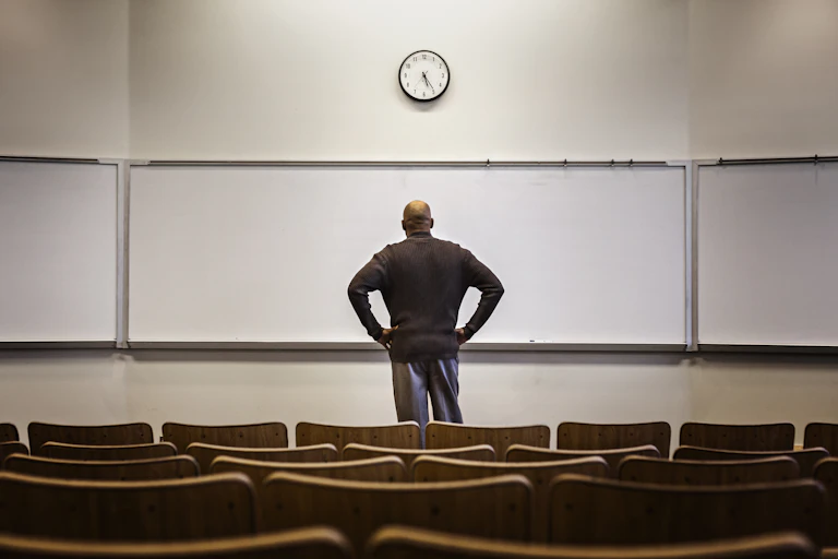 A man standing at a blank whiteboard, in a classroom.