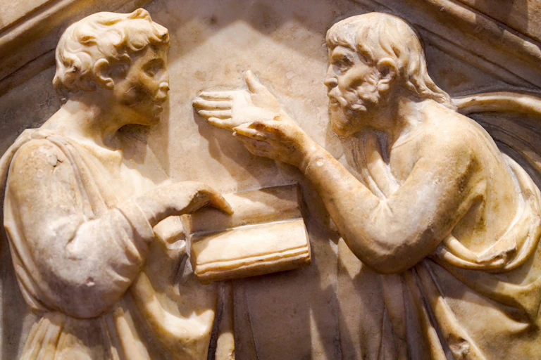 Relieve de un hombre hablando con las manos extendidas a otro que señala una cosa en un libro.