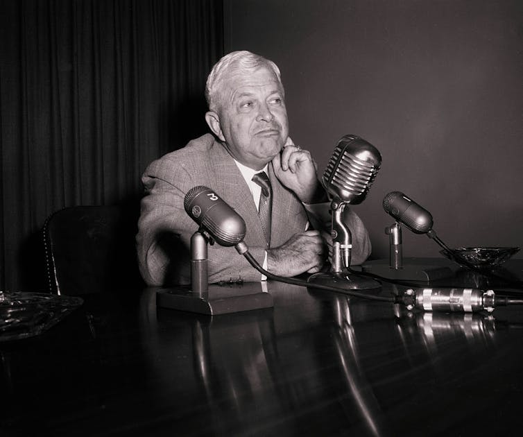 A black and white photo of a man with white hair, wearing a suit and tie and sitting at a table with microphones.