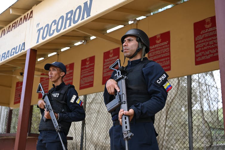 Two police officers wearing bulletproof vests and holding large firearms stand under yellow sign that says 'Tocoron'