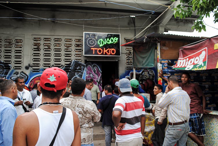 Over a dozen men stand outside a modest building that has a neon sign that says 'Disco Tokio'