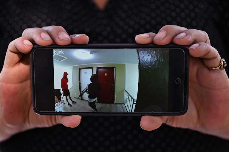 Two hands holding a mobile phone displaying an image of armed men in a residential hallway