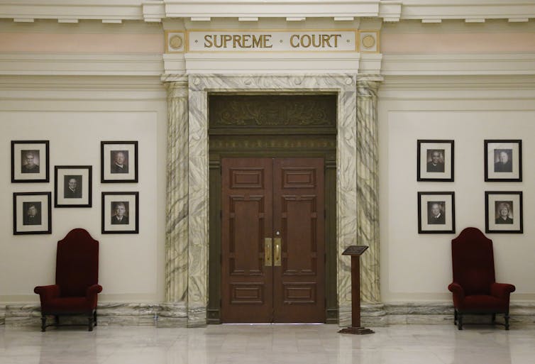 Tall, carved wooden doors in a white-walled hallway, with two chairs framing the door and photos on the walls.
