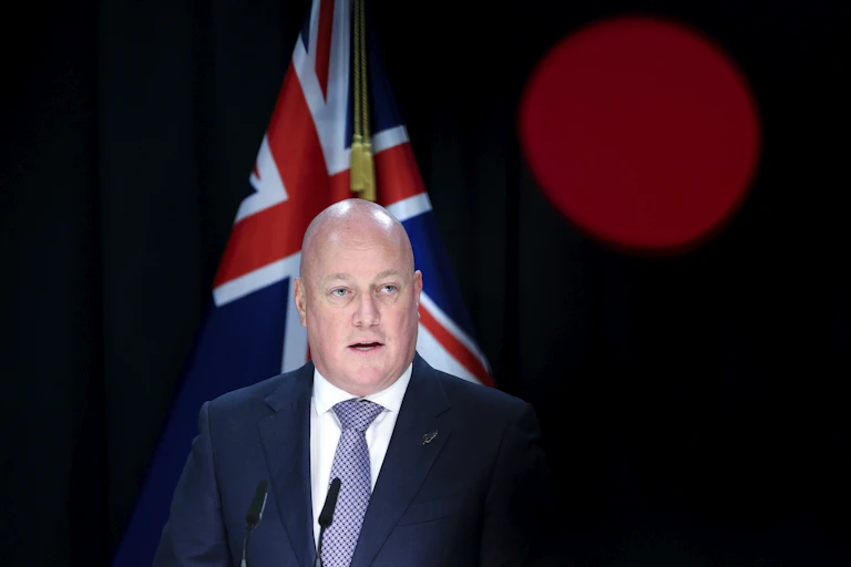 Christopher Luxon standing in front of a New Zealand flag and a dark background