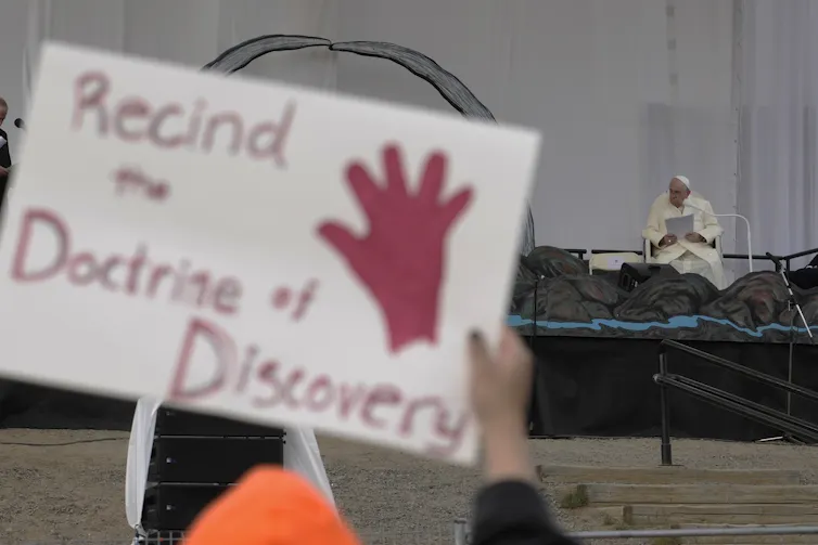 A sign that says 'rescind the doctrine of discovery' as a man in white robes sits on a stage.