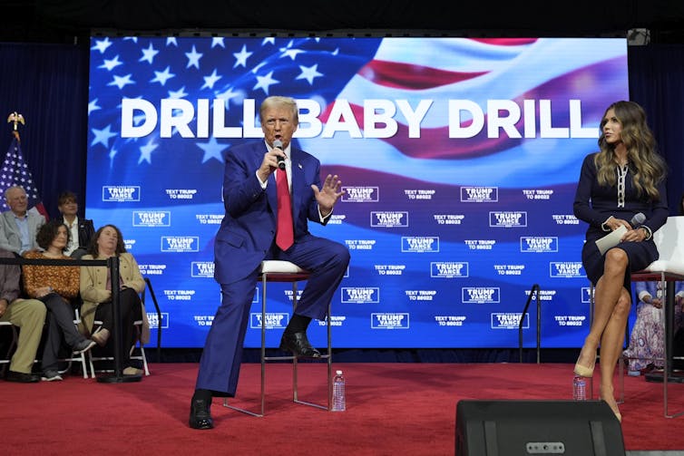 Donald Trump speaks into a microphone while sitting in front of a big screen that reads
