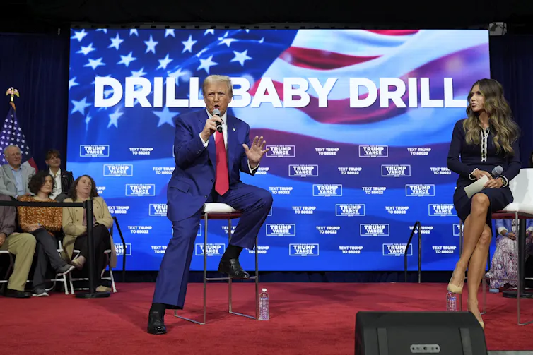 Donald Trump speaks into a microphone while sitting in front of a big screen that reads