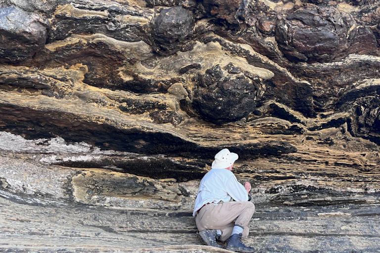 A man on his haunches on a rocky outcrop, looking at dark striations beneath a prominent dark protrusion