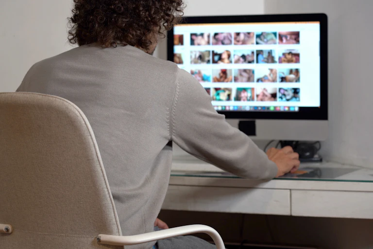 View from behind of a man looking at an adult website, with out of focus thumbnails of pornographic material