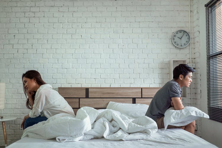 A young couple sitting on opposite sides of the bed, back to back, not speaking