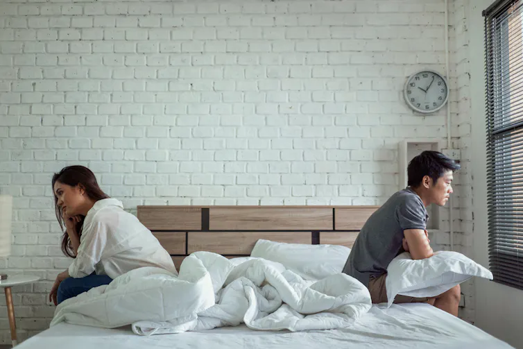 A young couple sitting on opposite sides of the bed, back to back, not speaking