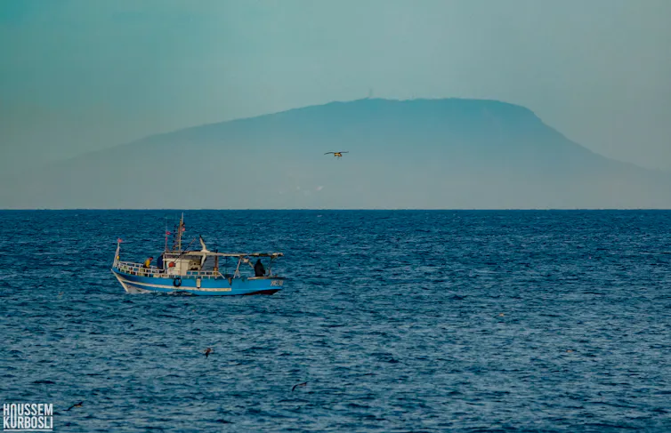 A small light blue fishing boat on a calm blue sea, an island visible behind it