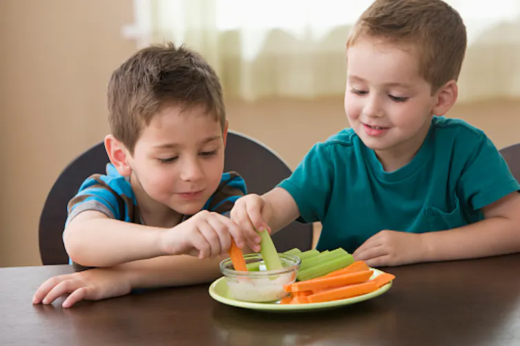Two boys dip vegetables into a bowl of ranch dressing.