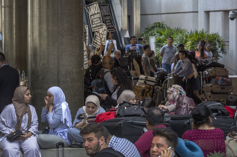 Women and children sit with their baggage in an airport as a protest goes on in the background
