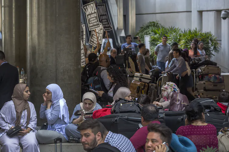 Women and children sit with their baggage in an airport as a protest goes on in the background