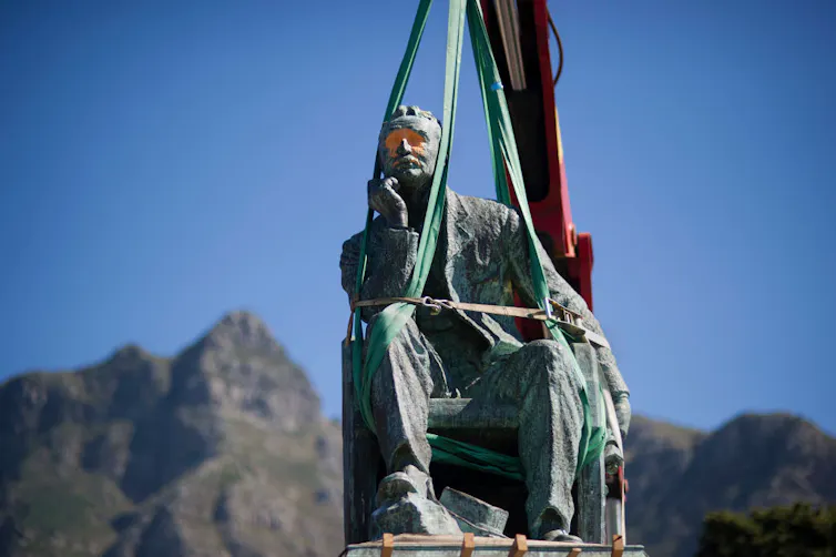 A statue of a seated man in a suit resting his head on one hand is hoisted into the air by a crane, the man's eyes smudged with red paint.