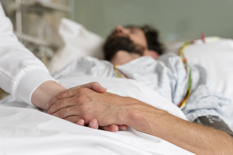 A person with eyes closed lies in bed with a tube attached to his body while another clasps his hand.