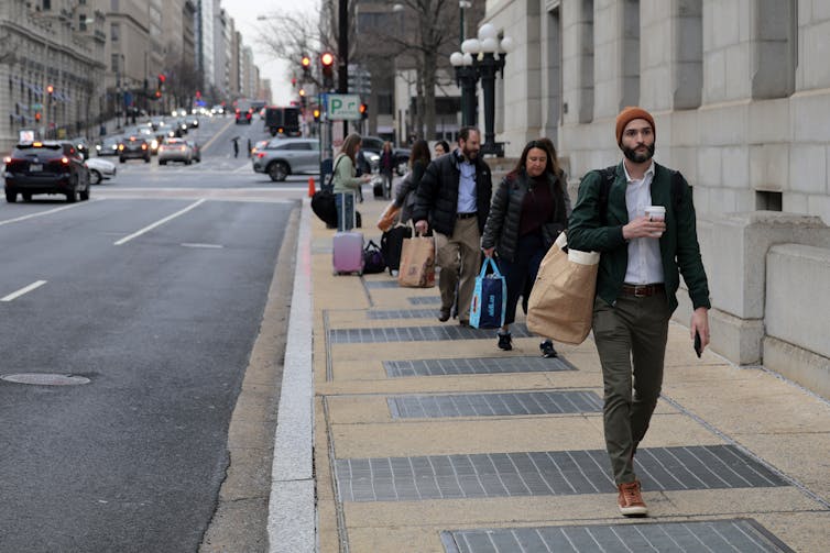 A row of people walk holding bags on a sidewalk in a city street.