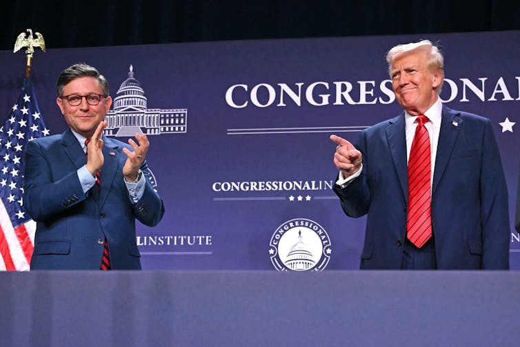 Two white men wear navy suits and stand at a podium in front of a blue backdrop and American flag.