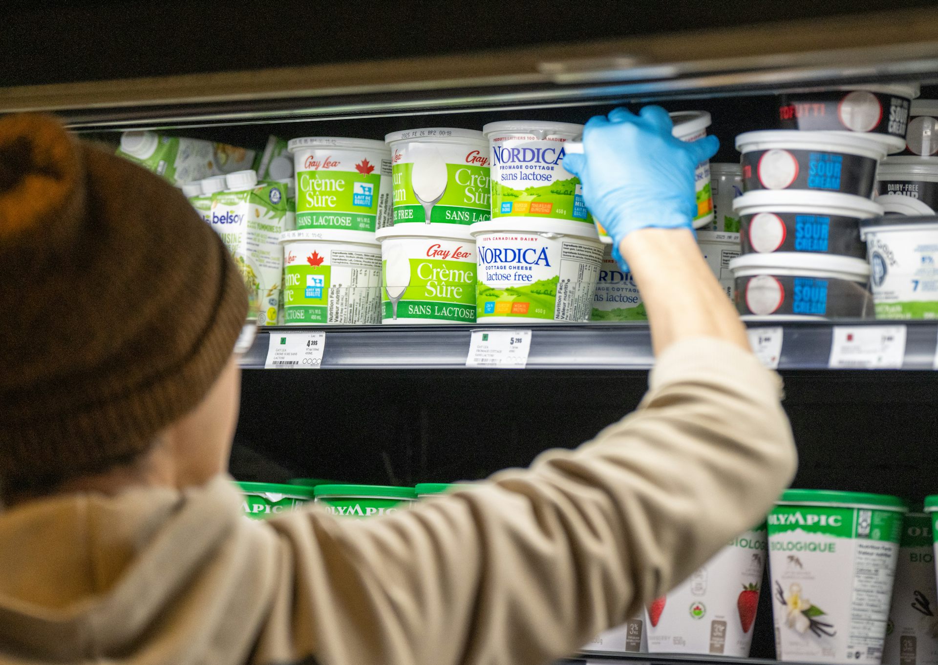 A person, seen from behind, places a container of sour cream on a refrigerated shelf in a grocery store