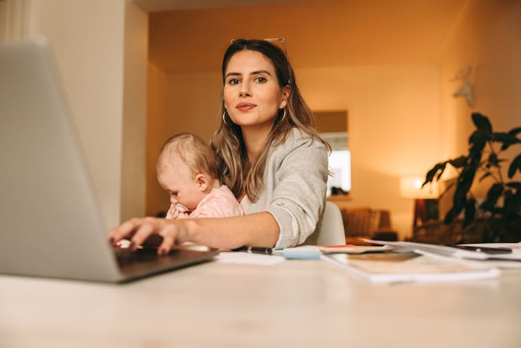 Young woman with shoulder length hair sitting at a desk typing, with a baby on her lap.