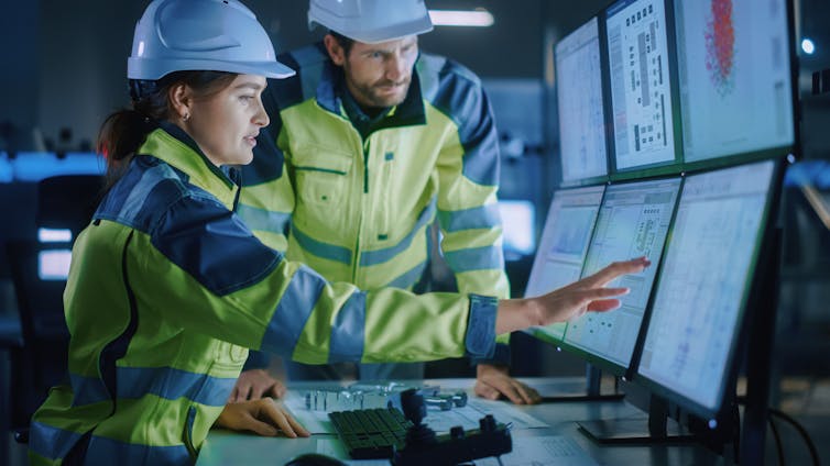 A young women in hi viz jacket and hard helmet sitting at computer screens while a man in hi viz jacket and hard hat looks on