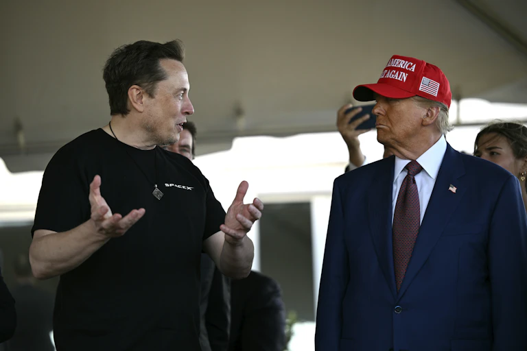 Two white men, one with brown hair and one wearing a red baseball cap that says Make America Great Again, converse