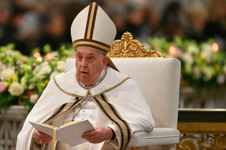 Un homme en chapeau et manteau à mitre blanche, assis sur une chaise blanche et dorée, tenant un document ou un livre