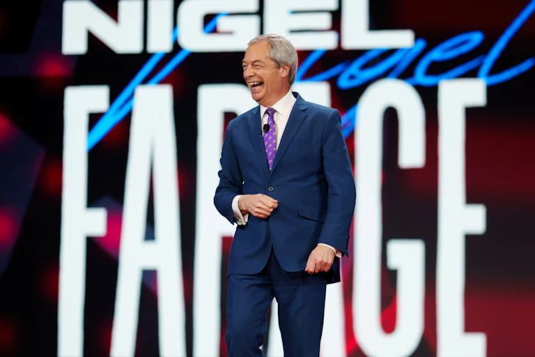 Nigel Farage smiling while walking on a stage, with his name in huge neon letters behind him