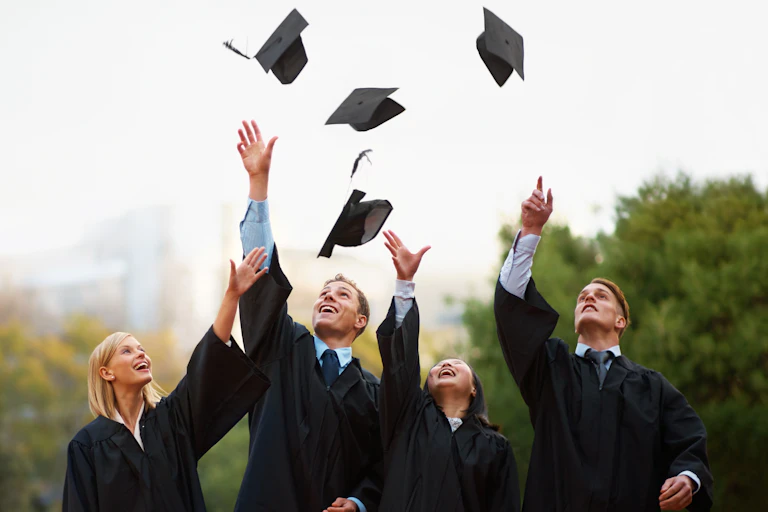 quatre jeunes diplômés (deux garçons et deux filles) en toges jettent leur toque en l'air.