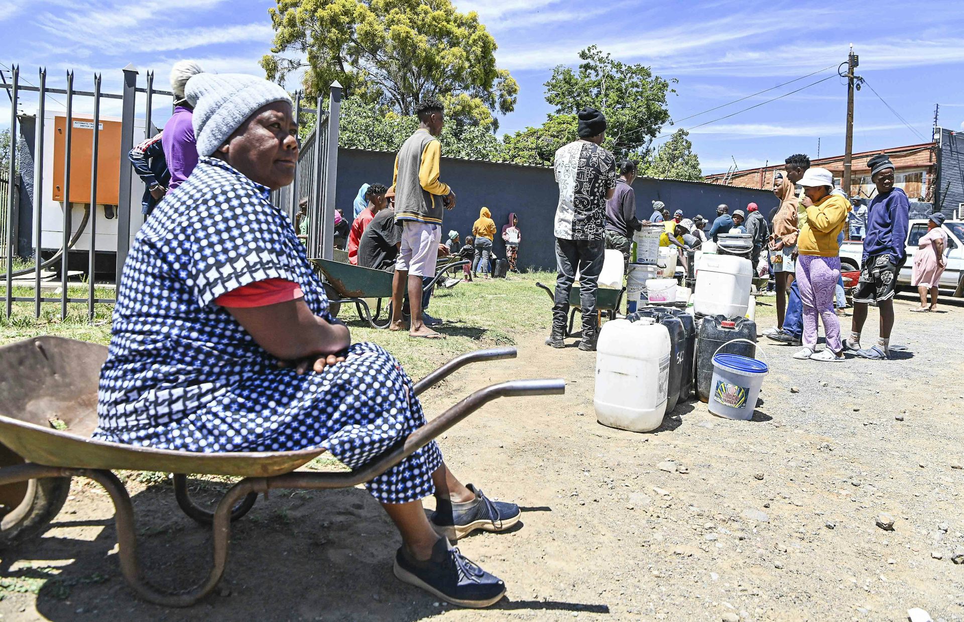 A woman sits on a wheelbarrow at the end of a queue of people lining up with buckets and containers for water