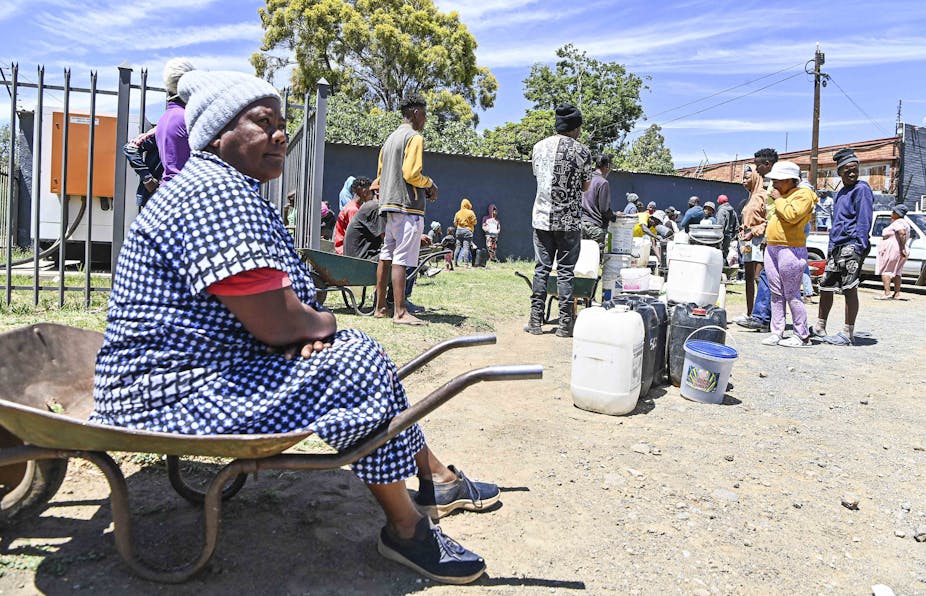 A woman sits on a wheelbarrow at the end of a queue of people lining up with buckets and containers for water