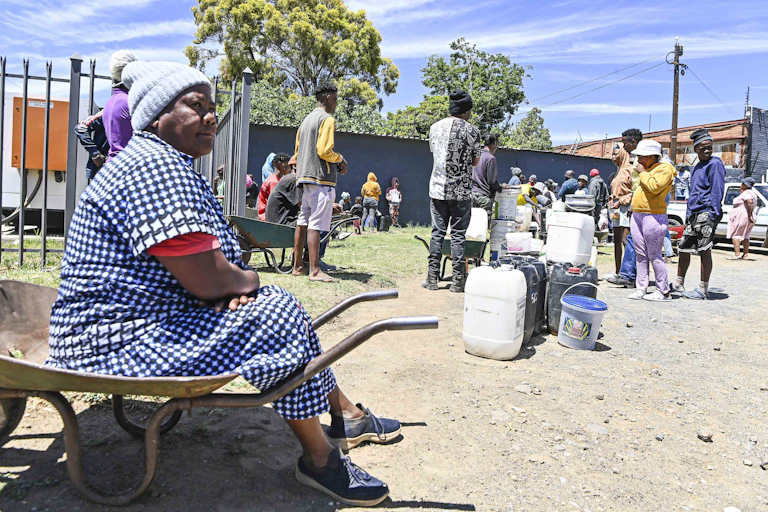 A woman sits on a wheelbarrow at the end of a queue of people lining up with buckets and containers for water