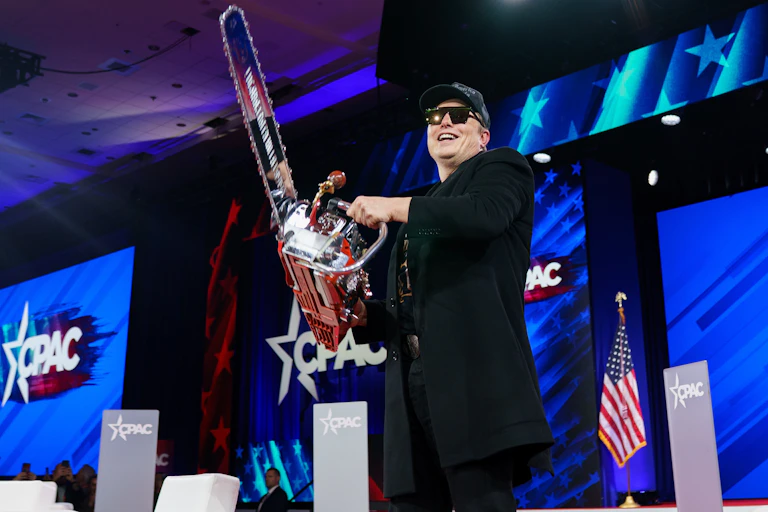 Elon Musk on a stage holding a chainsaw.