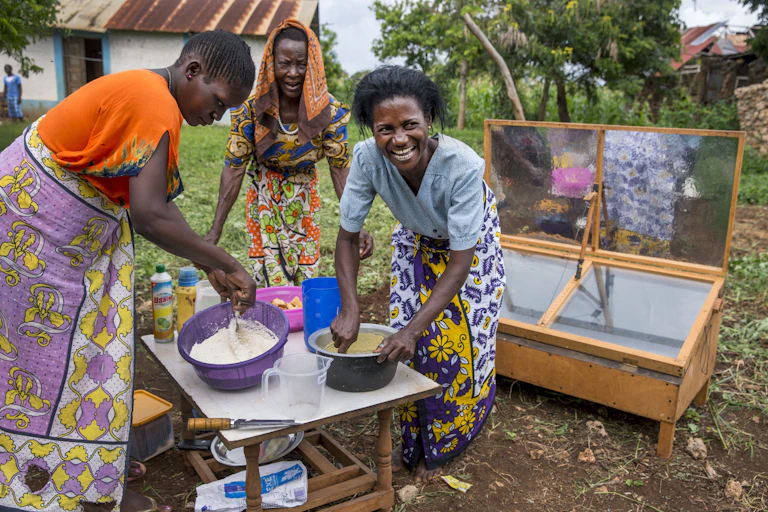 Three women mix ingredients in two bowls on a table outdoors, ready to put a dish into a solar oven for cooking