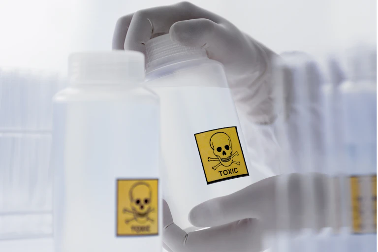 Gloved hands holding bottle with clear liquid and a skull and cross bones sticker reading "TOXIC"