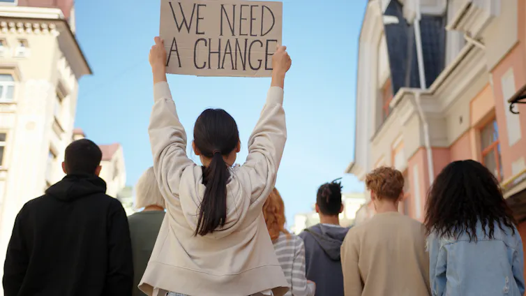 Young protesters seen from behind, with a young woman in a white shirt holding up a sign reading 'We need a change'