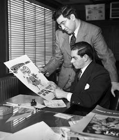 A black and white photo of two men in suits looking at a large illustration of a superhero.