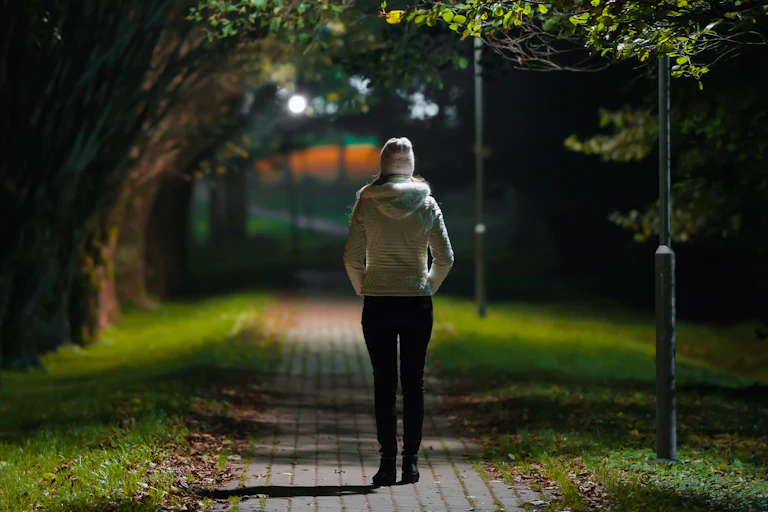 A young woman walking alone along a lit path in a park at night