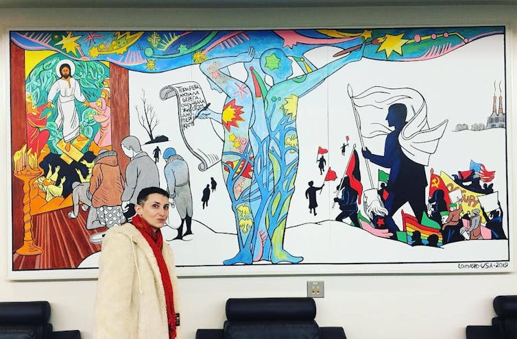 A woman with short brown hair stands in a white coat in front of a colorful indoor mural with two blue figures at the center.