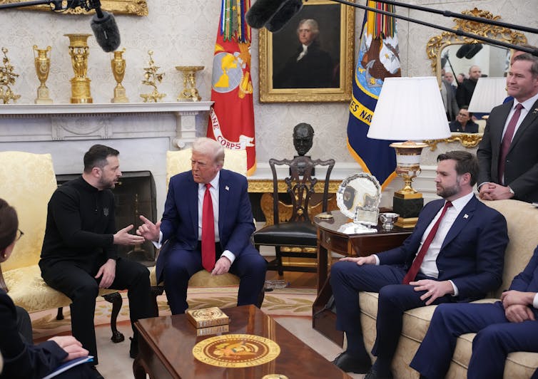Three men sit on couches and chairs around a wooden table, with microphones above them.
