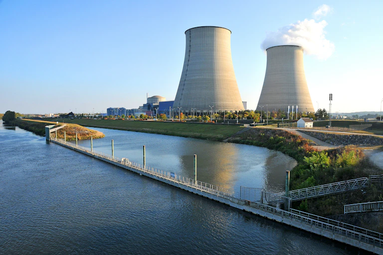 A nuclear power plant, with two large concrete towers, one of which is blowing smoke, sits next to a river.