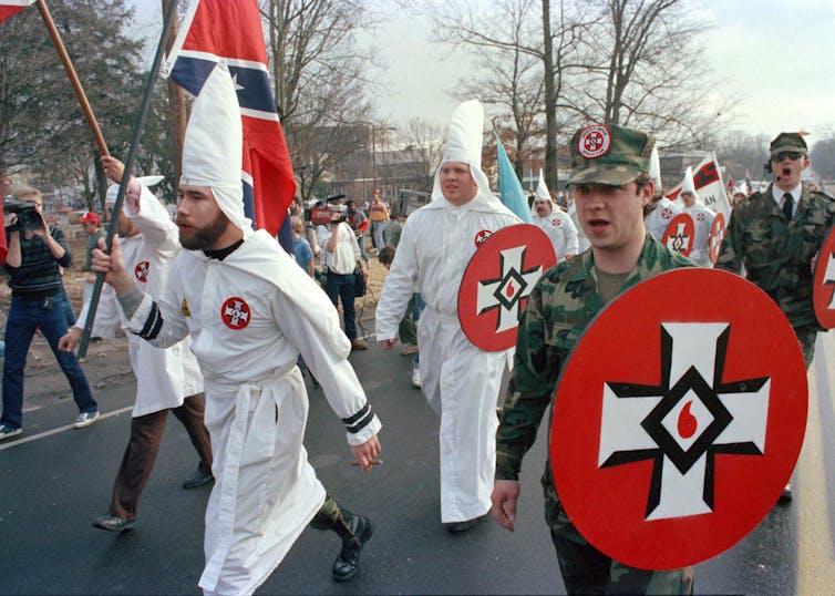 People in white robes and white pointed hats march down a street.