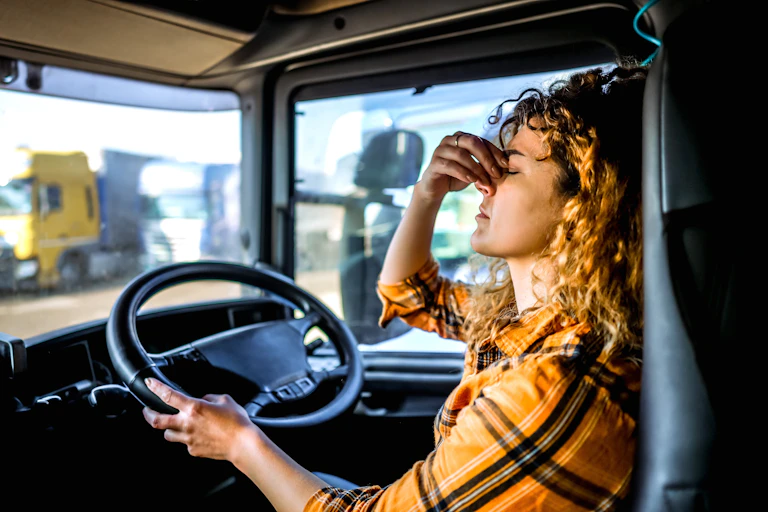 An exhausted female truck driver, eyes closed, sits in her truck.