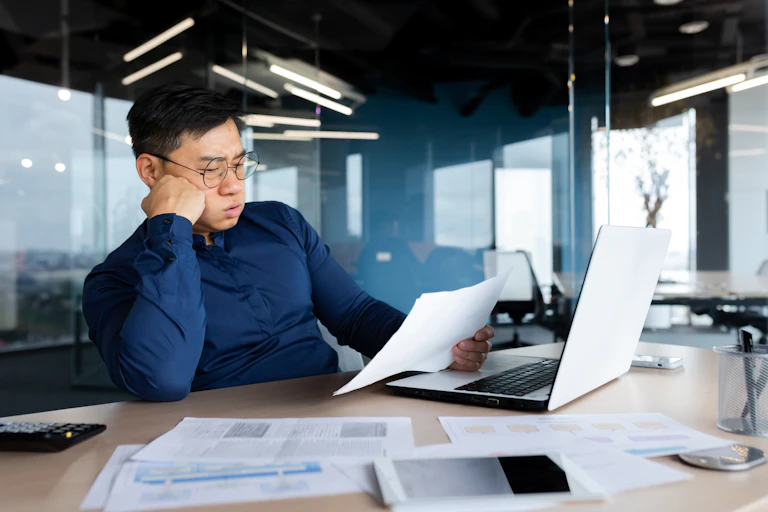 A frustrated man sits at a desk in front of a laptop looking at paper documents