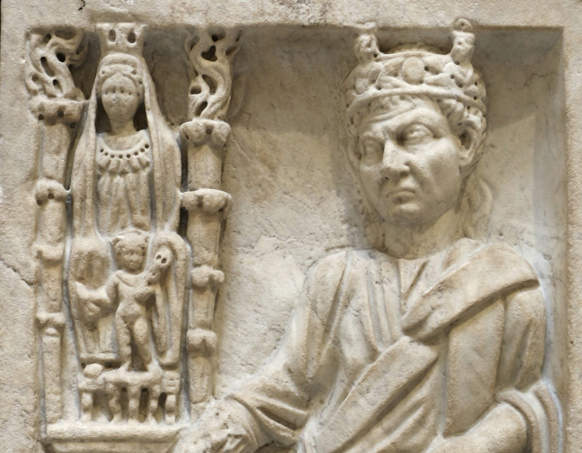 Um relevo de mármore com várias figuras gravadas: ao lado, uma coluna com uma mulher adornada e outra acompanhante. Ao lado deles está um homem com uma coroa, com uma das mãos em um pedestal.