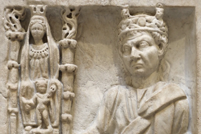 Um relevo de mármore com várias figuras gravadas: ao lado, uma coluna com uma mulher adornada e outra acompanhante. Ao lado deles está um homem com uma coroa, com uma das mãos em um pedestal.