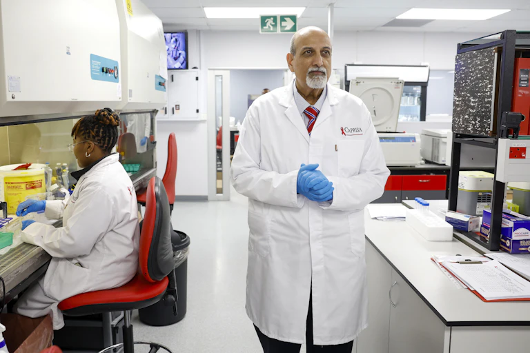 A man in a white lab coat, wearing blue surgical gloves, stands in a laboratory alongside a woman, similarly garbed, who is working on samples of some sort